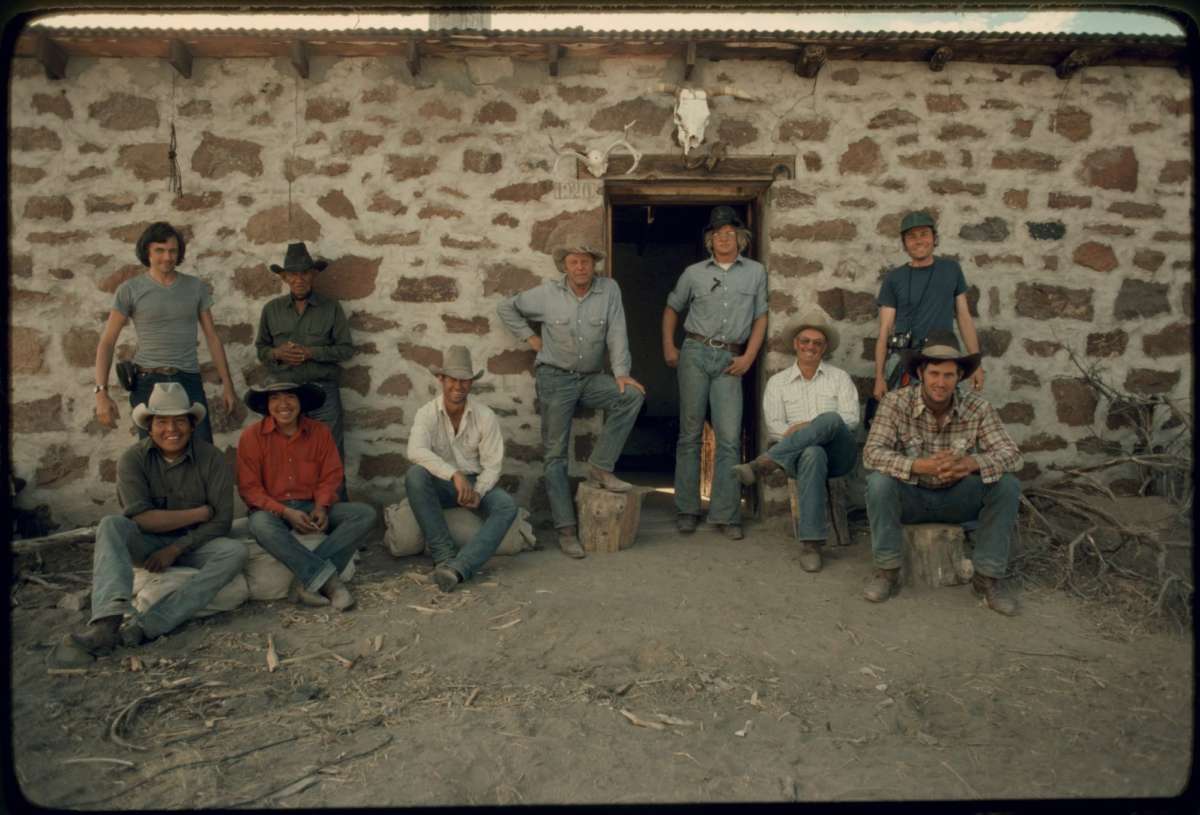 10 people wearing blue jeans and shirts, some seated on logs, some standing, all smiling in front of an old stone work cabin.